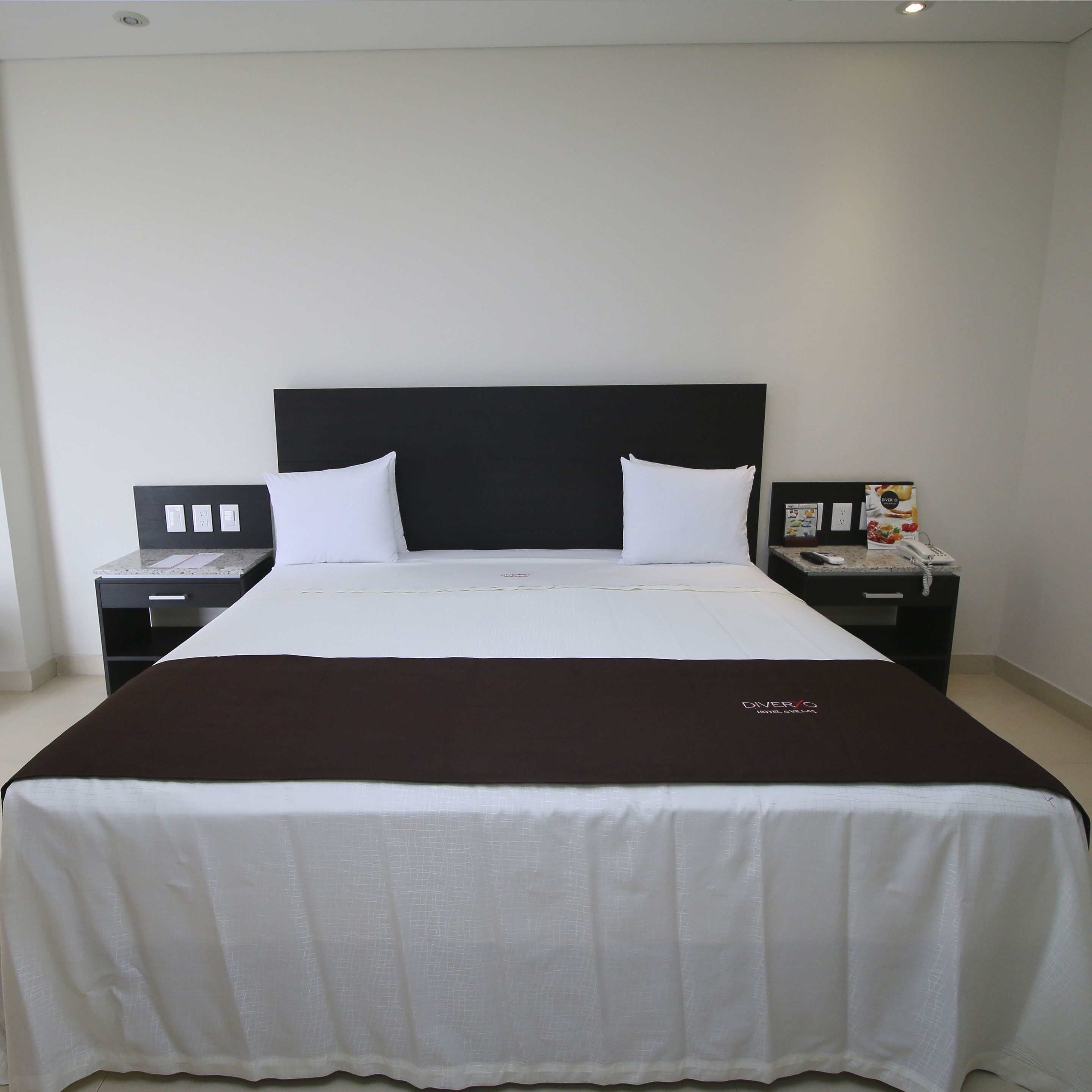 A hotel room with a large bed, two bedside tables, a black headboard, and a white bedspread.