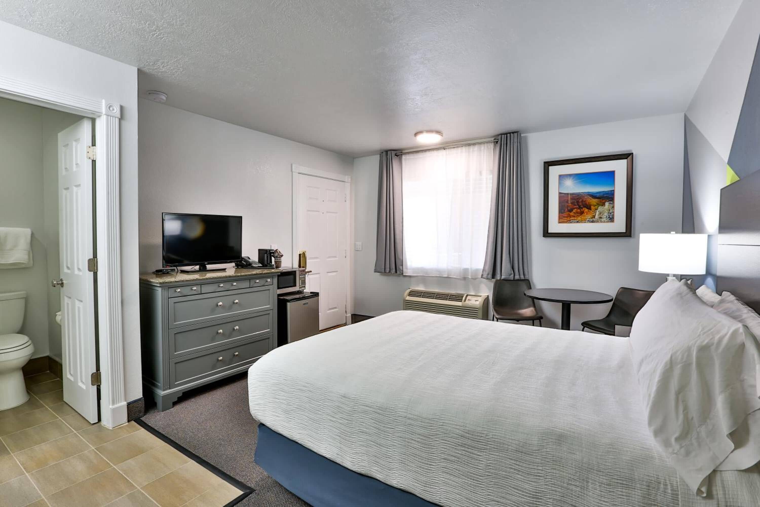 A hotel room with a large bed, a TV, a desk with a computer, a chair, a small table, and a window with curtains.