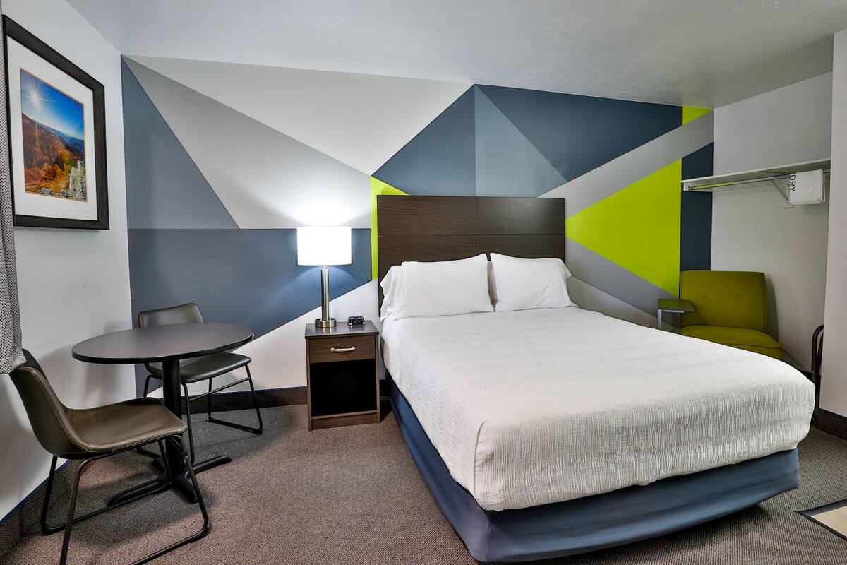 A hotel room with a bed, a desk, a chair, a nightstand, and a lamp.