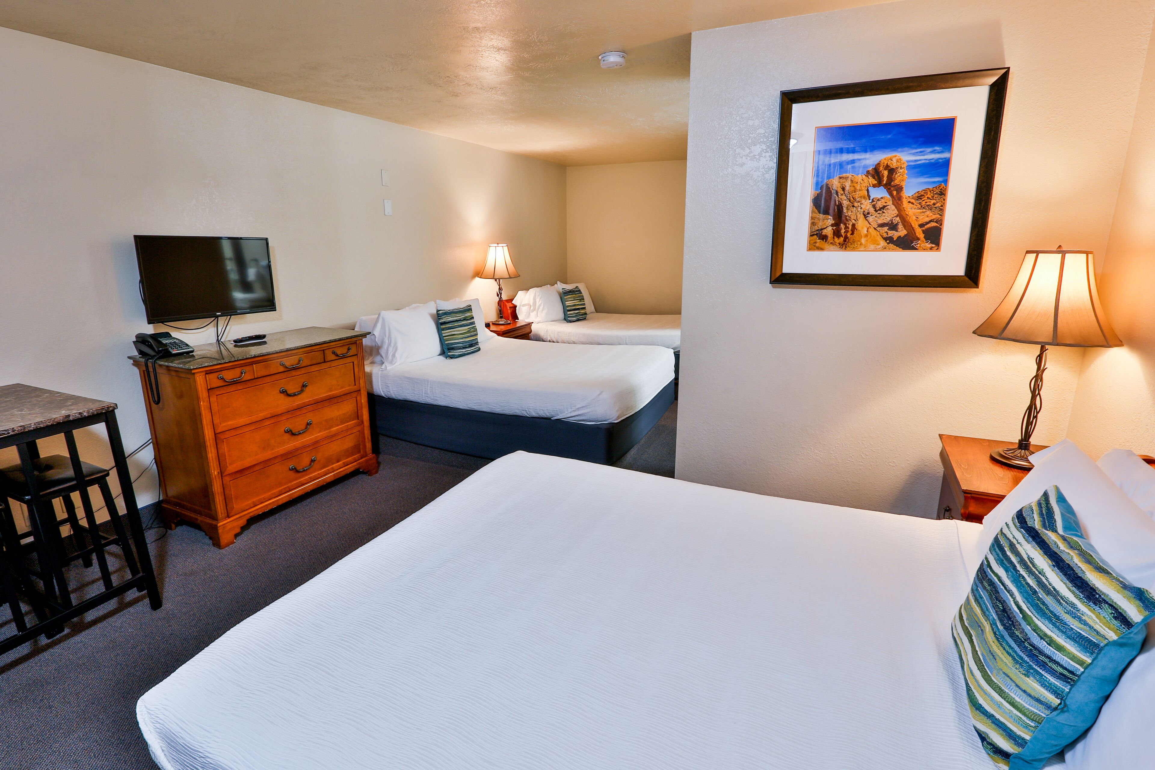 Une chambre d’hôtel avec un grand lit, un bureau avec une chaise, une commode, un téléviseur, deux lampes de chevet et un tableau encadré au mur.