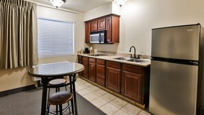 Cocina compacta con armarios de madera, un refrigerador de acero inoxidable, un microondas y una pequeña mesa de comedor con dos taburetes.