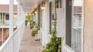 Un corredor exterior cubierto con varios ventanales, algunos con plantas, y una fila de luces fijadas a la pared.