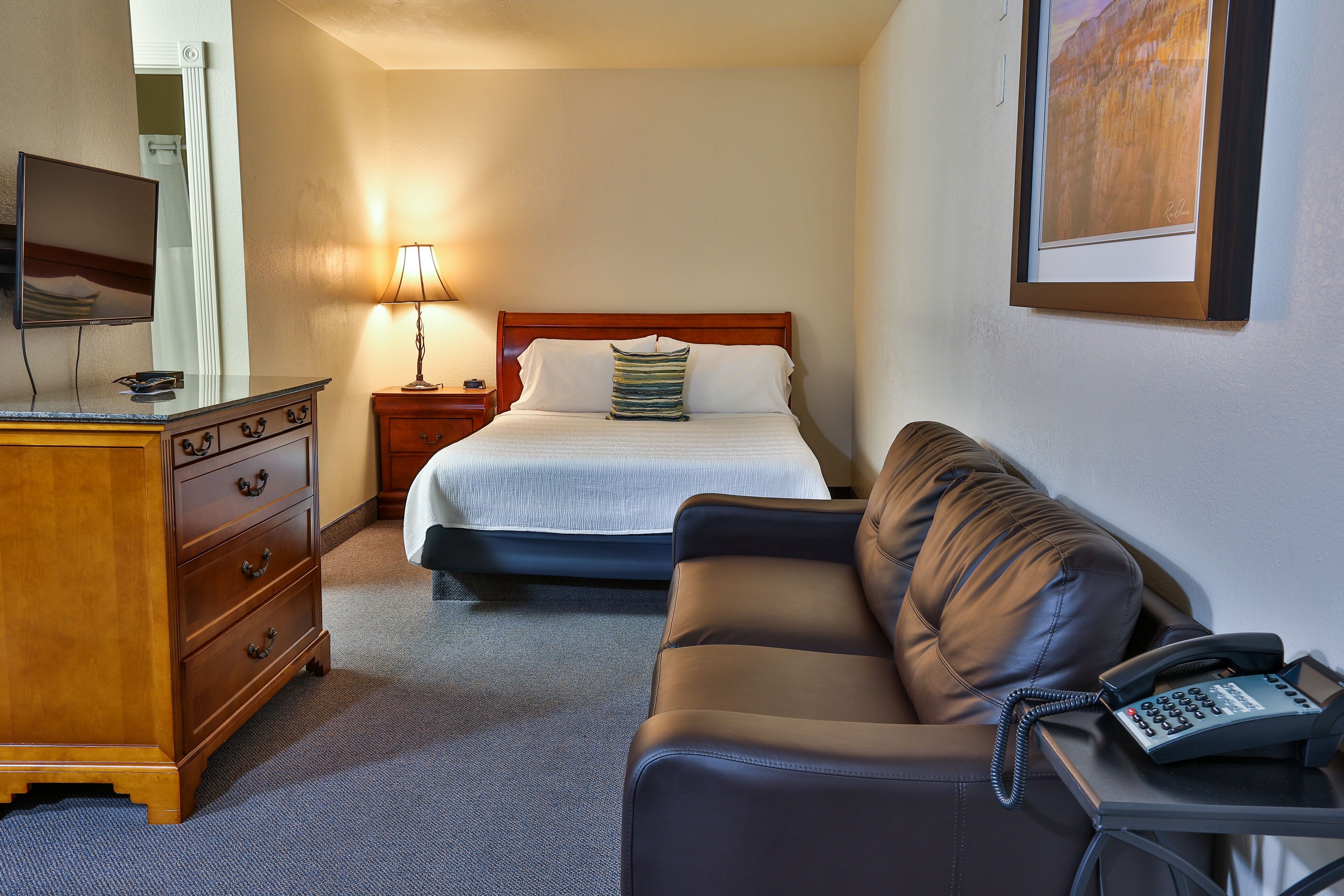 A hotel room with a bed, a wooden dresser, a leather sofa, a desk with a telephone, and a framed picture on the wall.