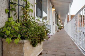 A covered outdoor corridor with a planter box filled with green plants and purple flowers, a white railing, and a light fixture on the ceiling. - Chalet Motel (St. George)