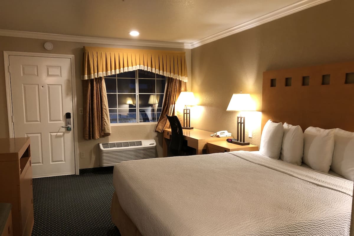 A hotel room with a large bed, a desk with a lamp, a chair, a window with curtains, and a door with a keypad lock.