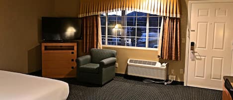 A hotel room with a bed, a chair, a TV, a heater, and a window with curtains.