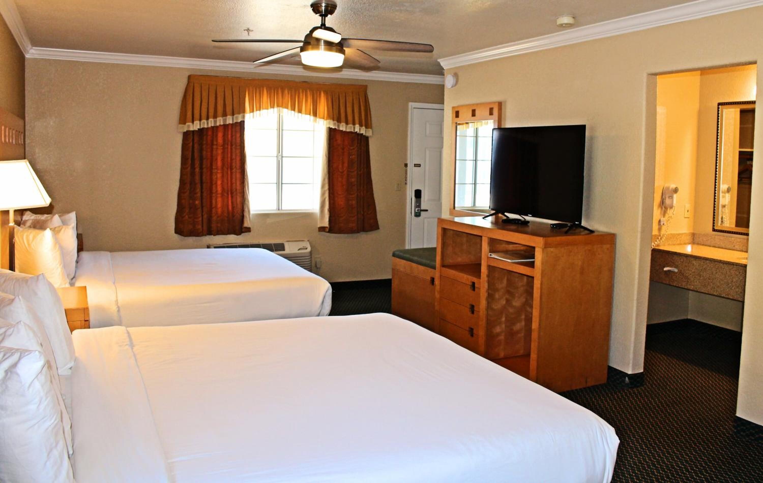 A hotel room with two beds, a television, a desk with drawers, a ceiling fan, and a window with curtains.