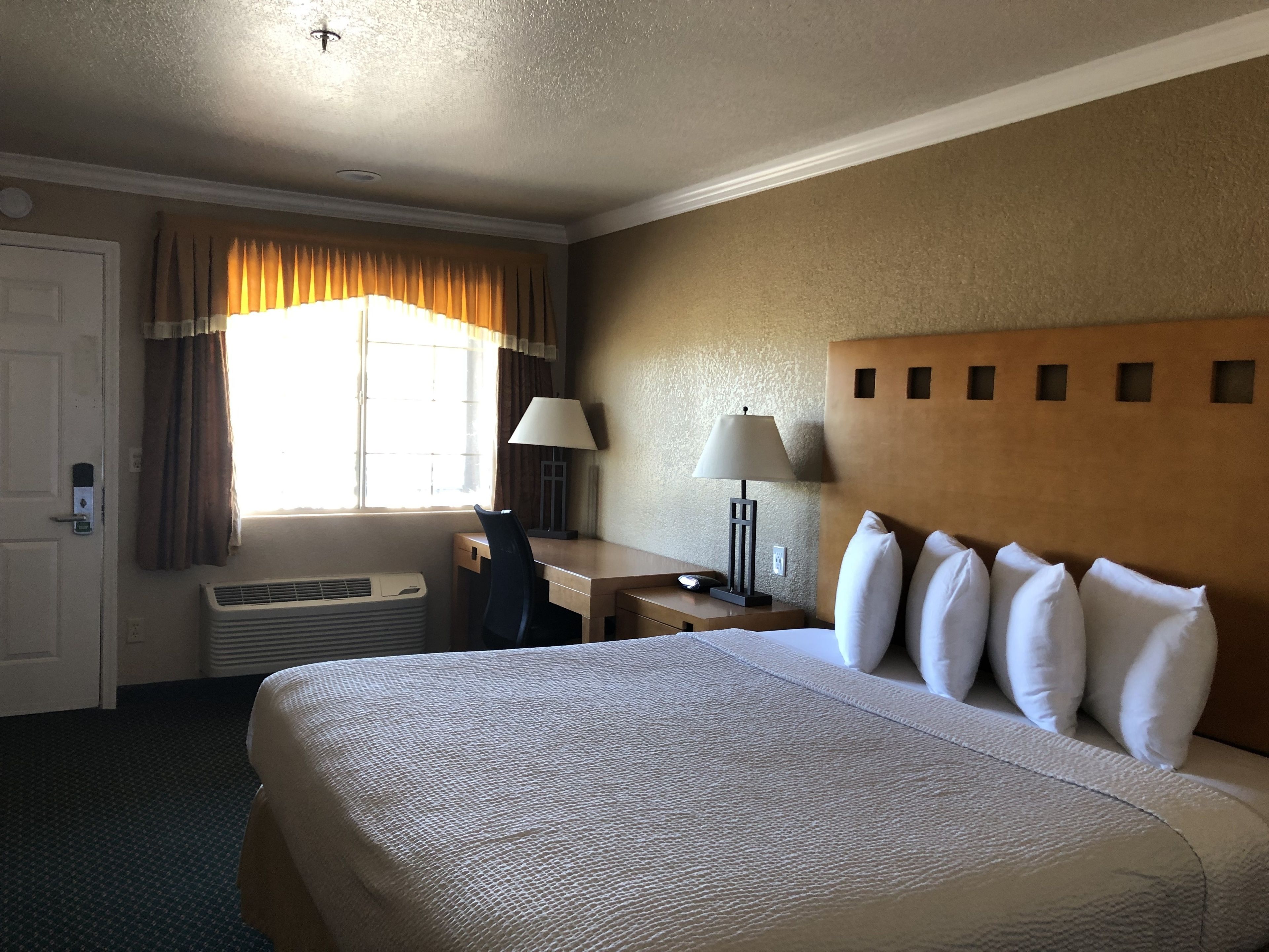 A hotel room with a large bed, a desk, a chair, a lamp, a window with curtains, and an air conditioning unit.