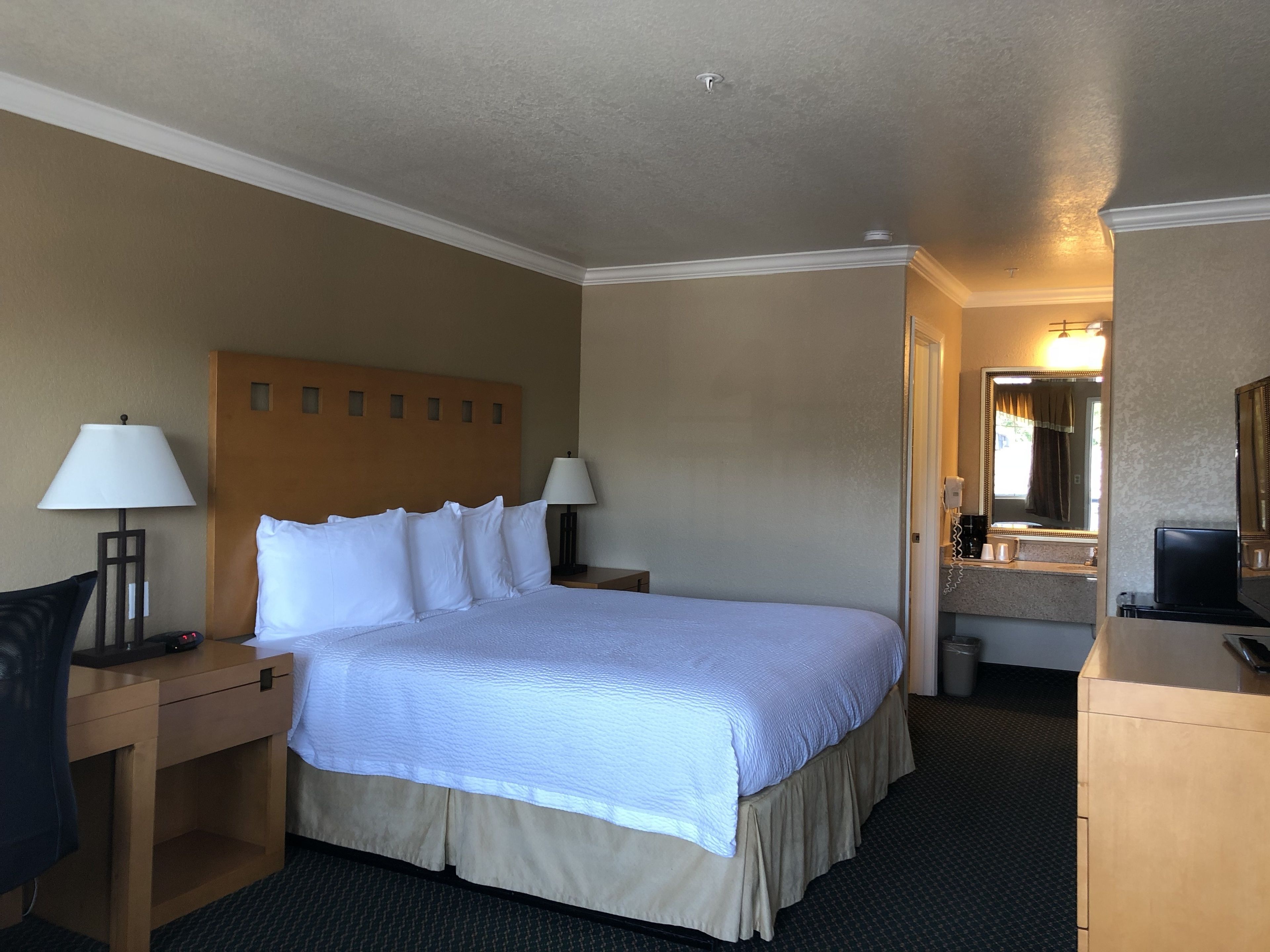 Habitación de hotel con cama, dos mesitas de noche con lámparas, escritorio con computadora, espejo y baño visible a través de una puerta abierta.
