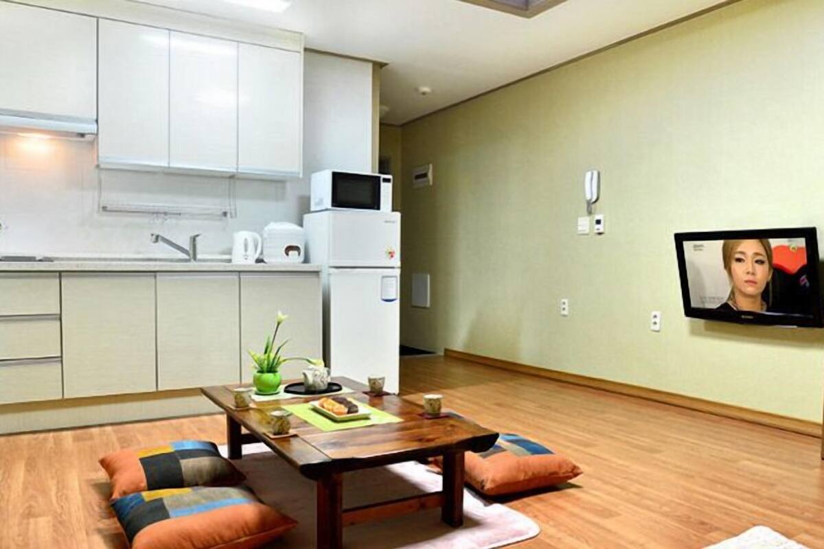 Un espace de vie compact comprenant un coin cuisine, un espace repas avec une table en bois et des coussins, et un téléviseur fixé au mur.