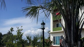 Un bâtiment moderne à la façade blanche et aux touches de vert, entouré d’une végétation tropicale et surplombé d’un ciel d’un bleu éclatant.
