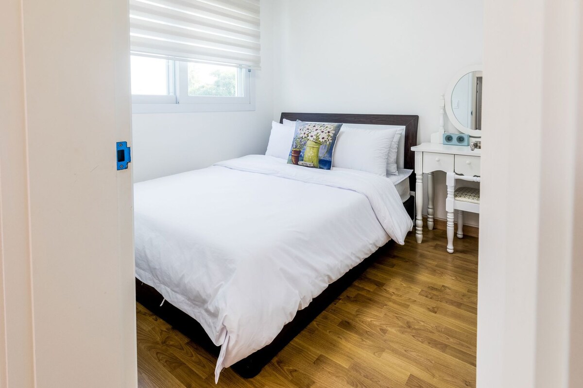 A bedroom with a bed, a bedside table with a mirror, and a window with blinds.