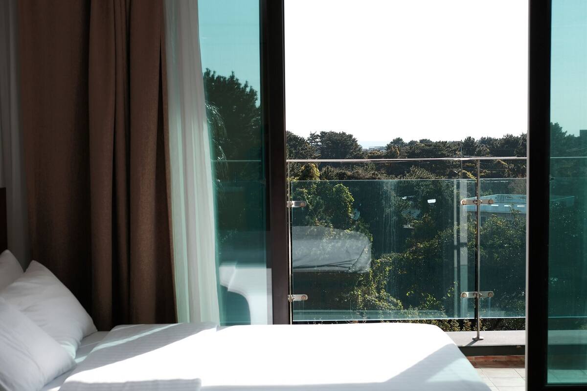 Een hotelkamer met een groot bed, bruine gordijnen en uitzicht op een balkon met groen en een heldere lucht.