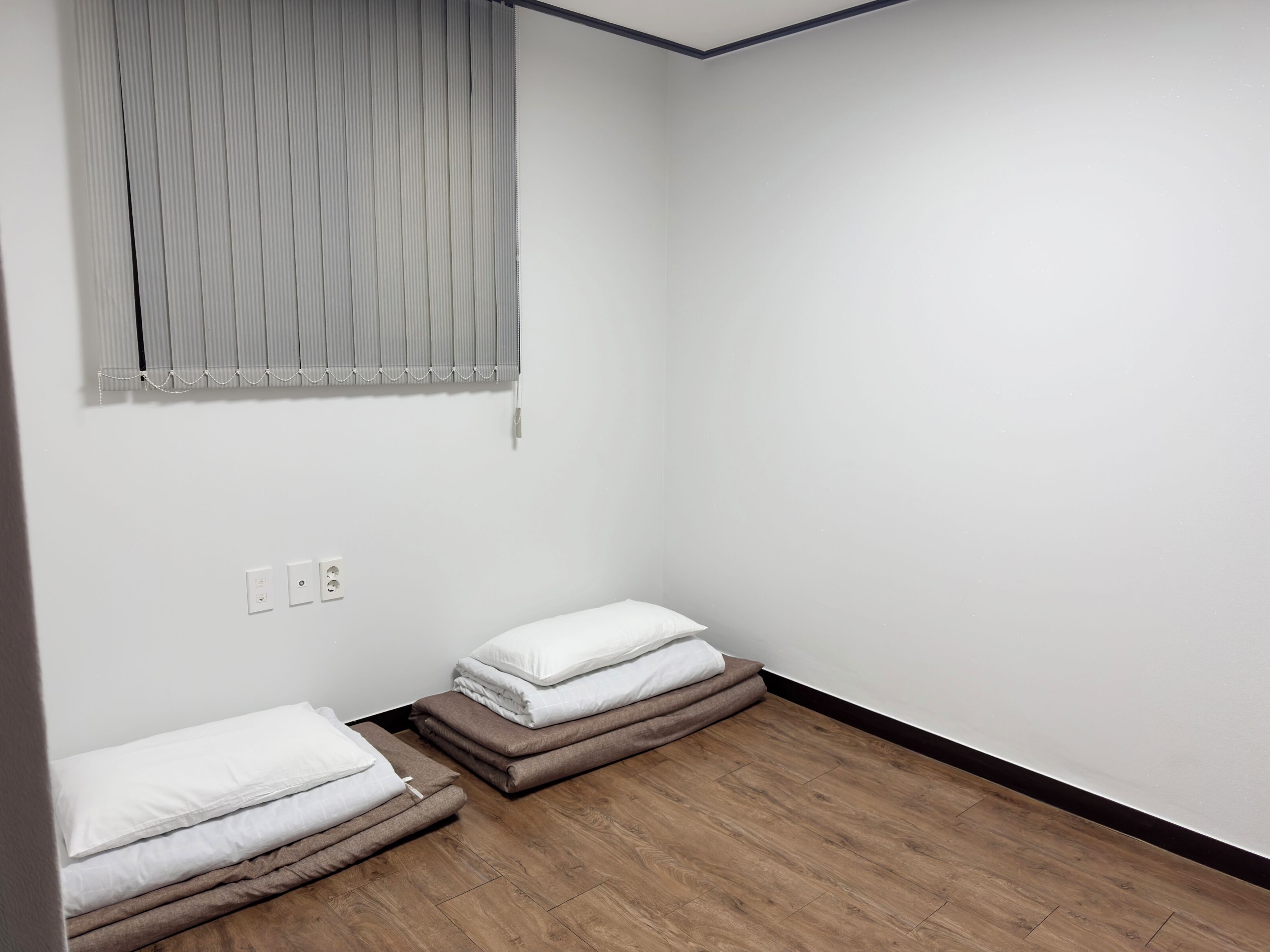 A small, empty room with a wooden floor, a window with blinds, and two beds with white and brown bedding.