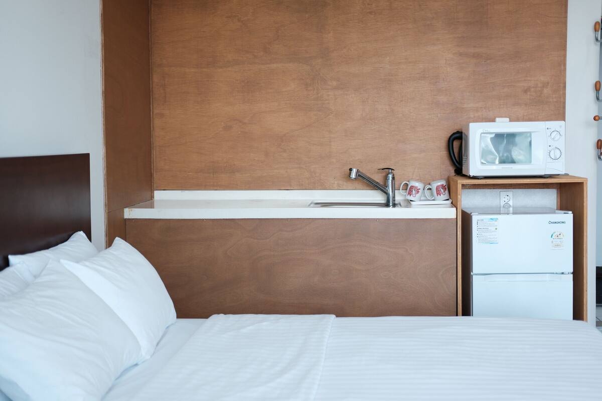 Een hotelkamer met een houten hoofdeinde, een bed met witte lakens, een witte wastafel met kraan, een magnetron op een plank, en een kleine koelkast.
