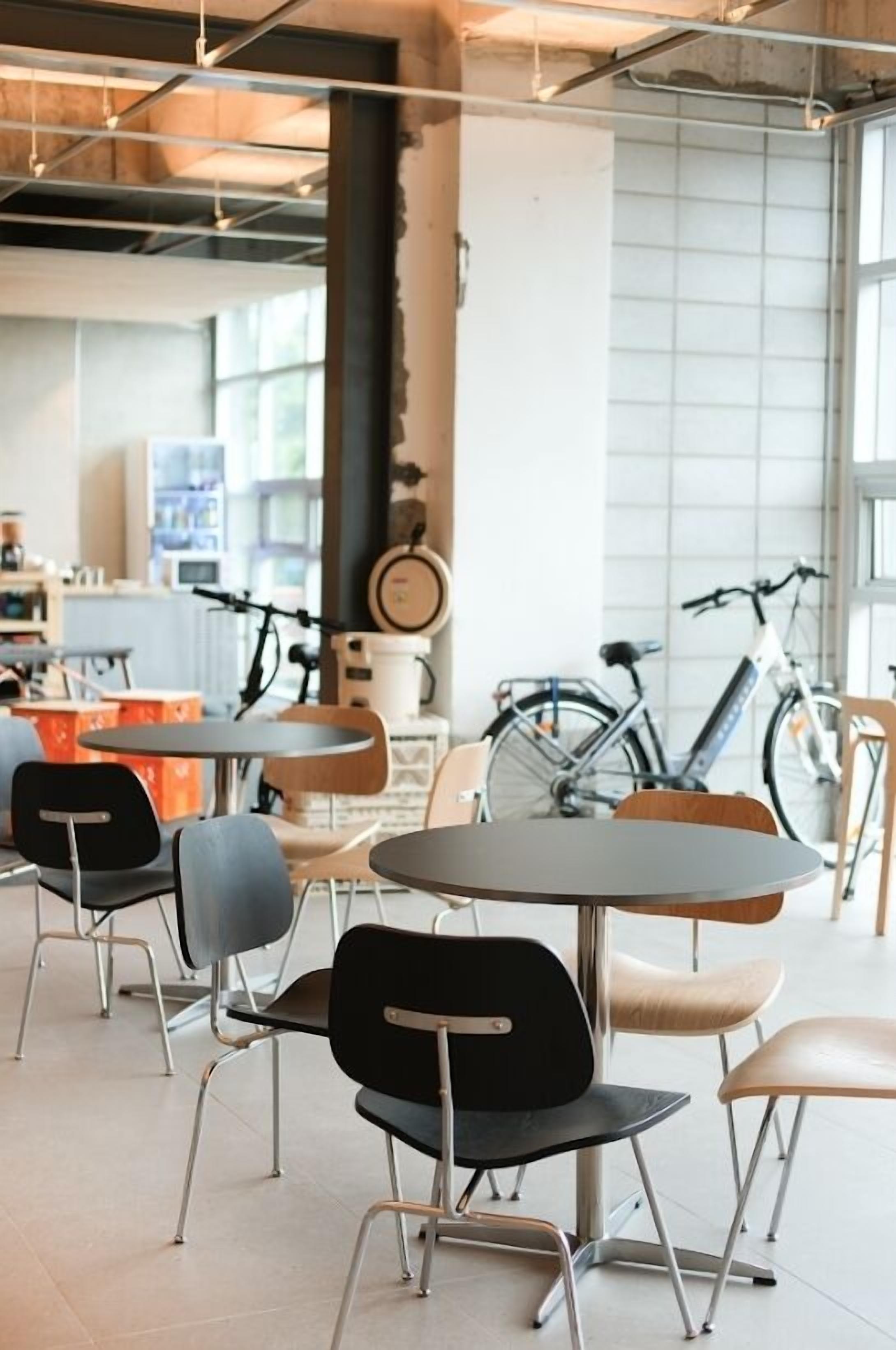 A modern cafe with round tables and chairs, bicycles in the background, and a visible kitchen area.