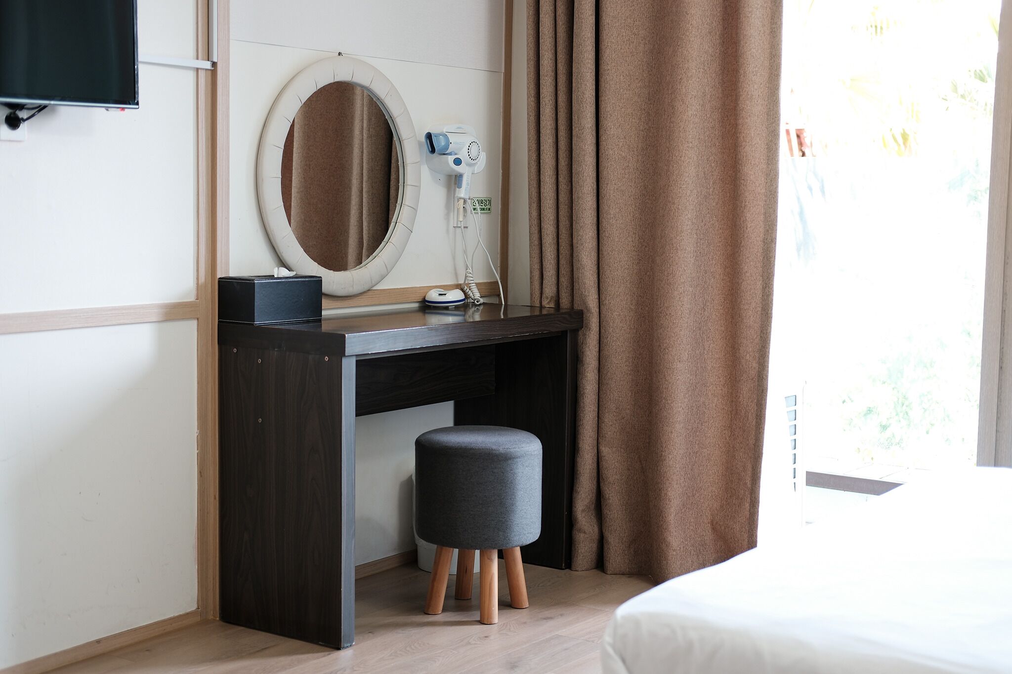 A hotel room with a dark wooden desk, a round mirror, a hairdryer, and a small grey ottoman.