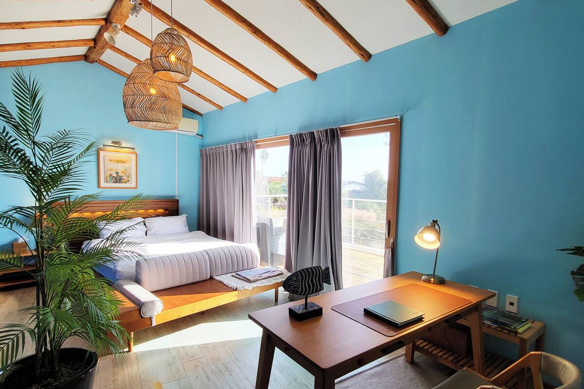 Une pièce comprenant un lit, un bureau, une chaise, un luminaire suspendu et une fenêtre avec des rideaux.