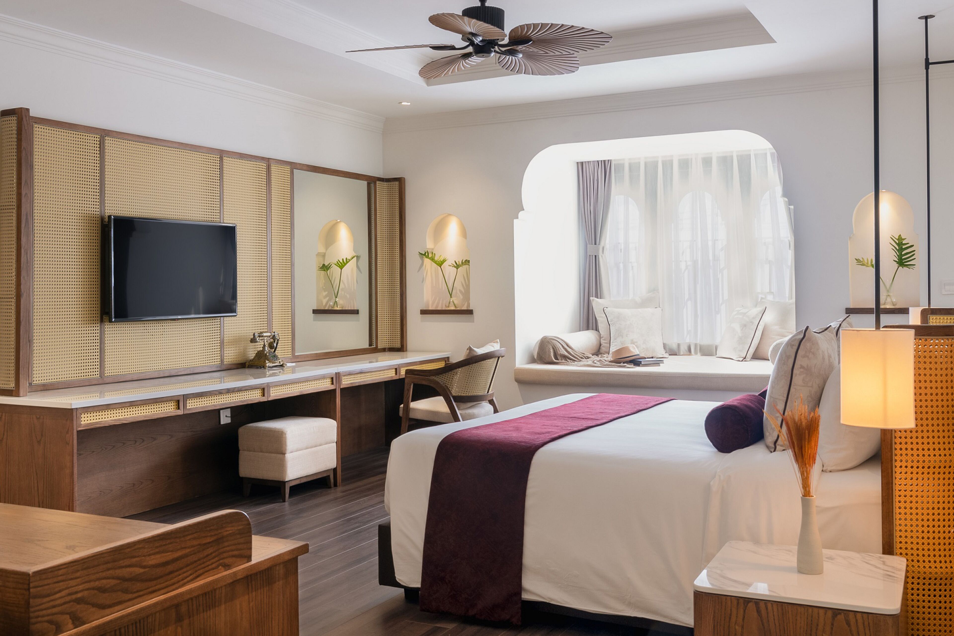A hotel room with a bed, a desk with a flat-screen TV, a seating area with a chair, a window with sheer curtains, and a ceiling fan.