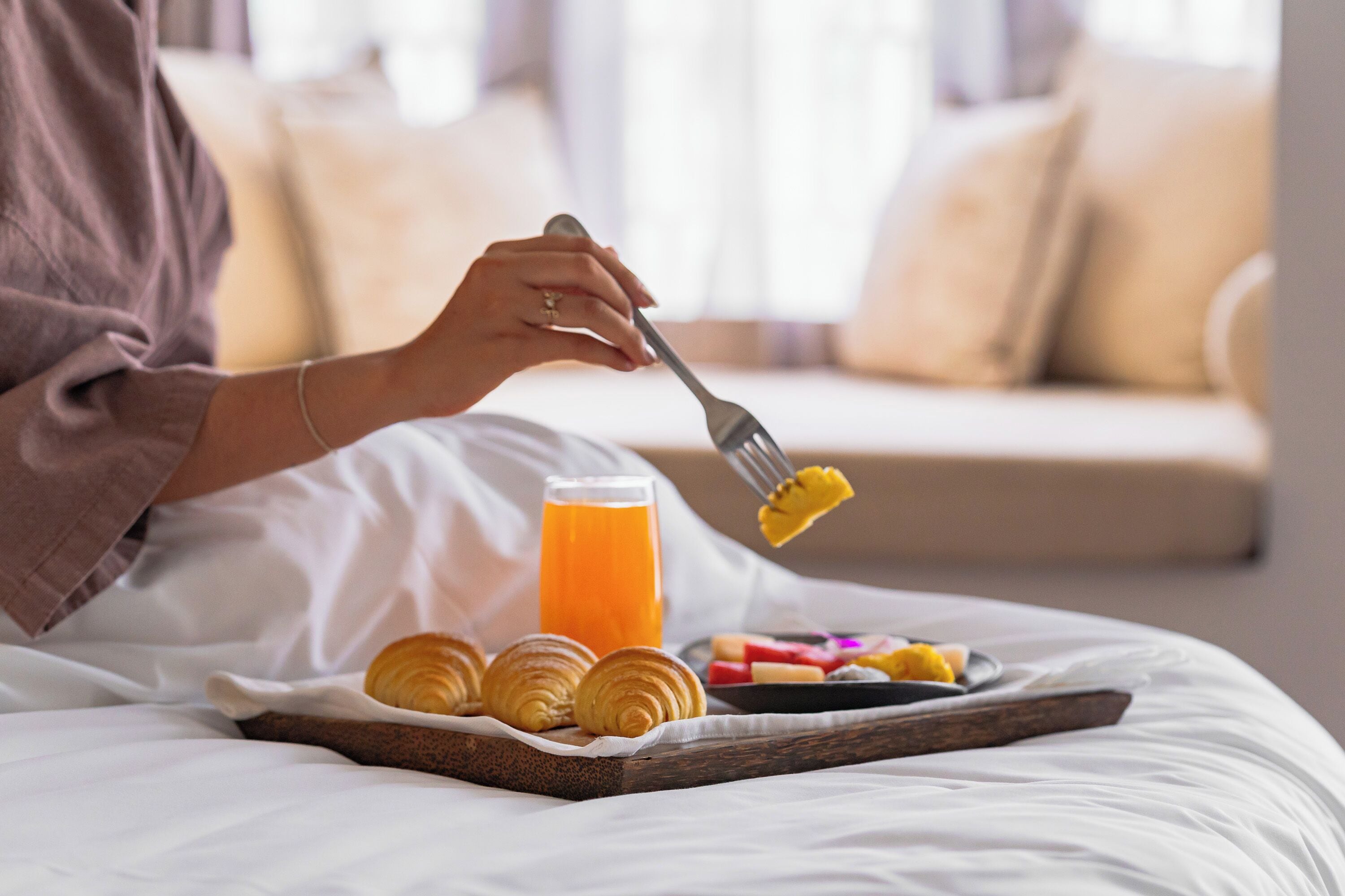 A person sitting on a bed with a tray of croissants, a glass of orange juice, and a plate of fruit.