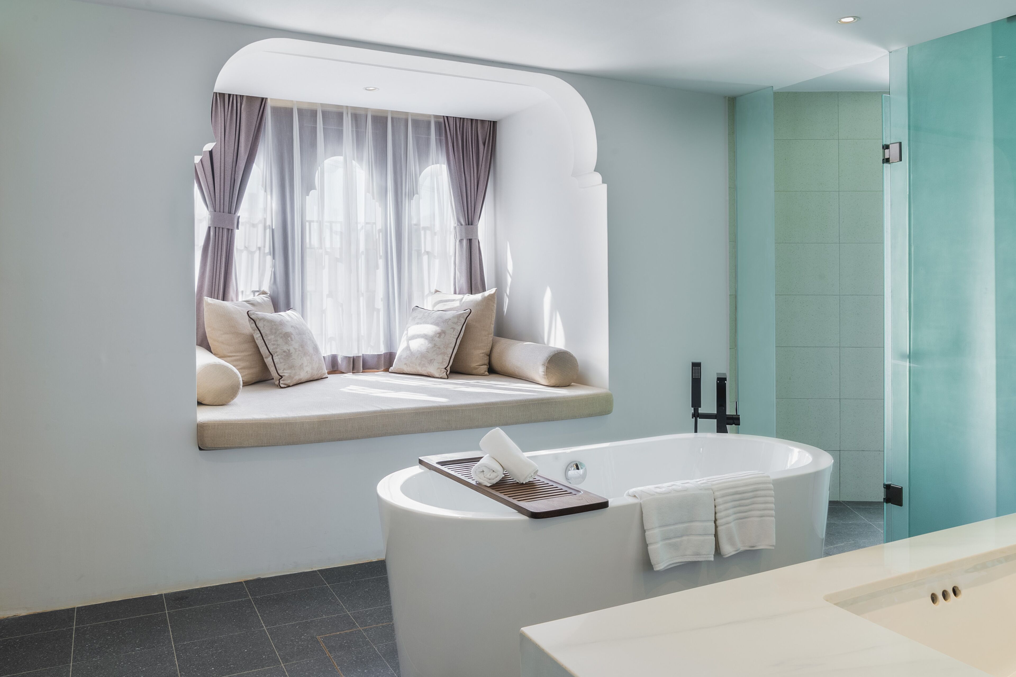 A modern bathroom with a freestanding bathtub, a window seat with cushions, and a glass shower enclosure.
