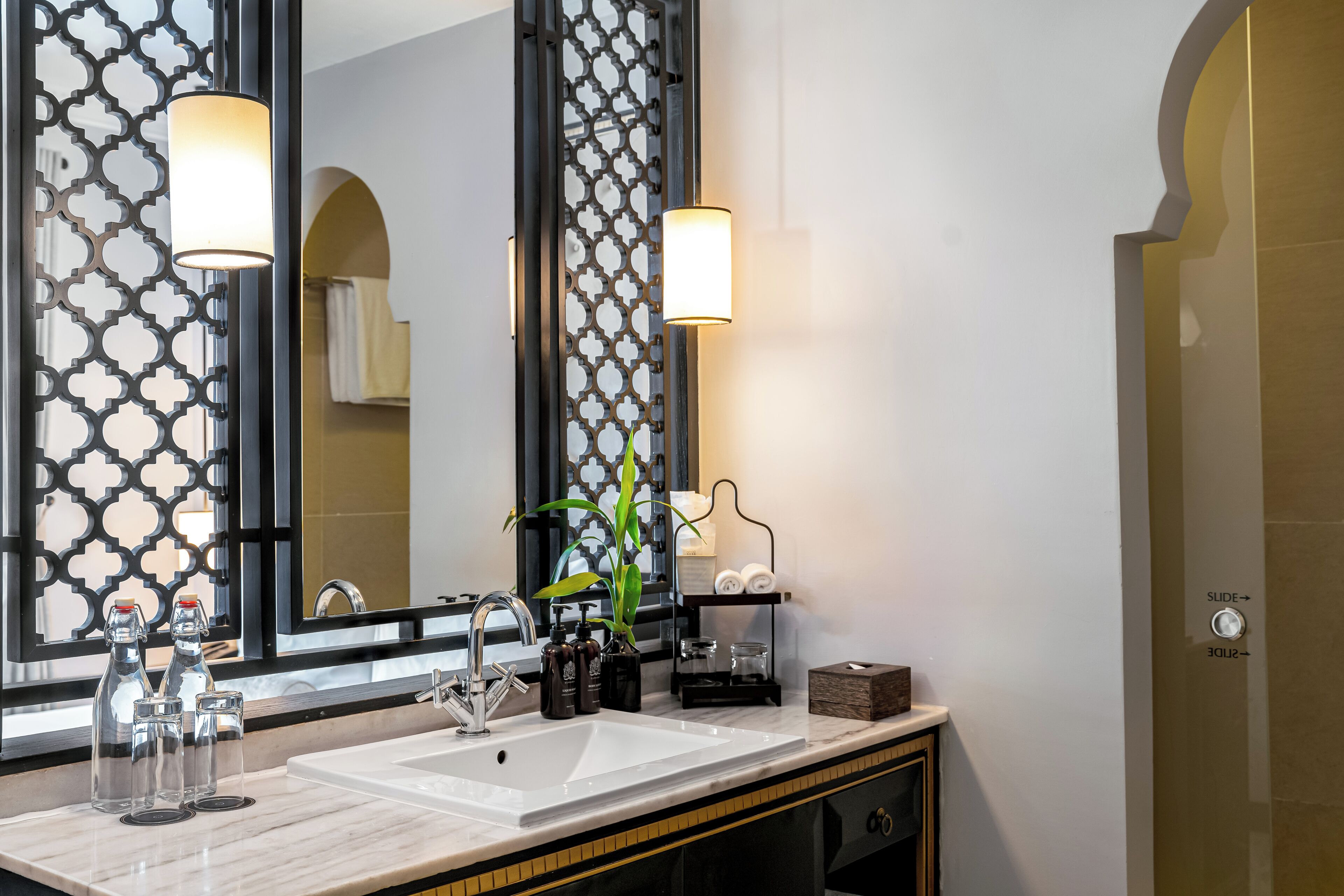Un baño con un lavabo blanco, dos llaves, un espejo con marco decorativo y una planta en maceta.
