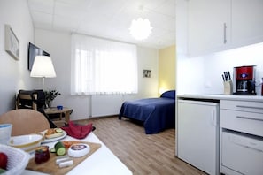 Habitación de hotel compacta con cama, una kitchenette pequeña, zona de comedor con productos para el desayuno y ventana con cortinas traslúcidas.