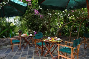 An outdoor dining area with wooden chairs and tables, covered by green umbrellas, surrounded by lush greenery and flowering plants.
