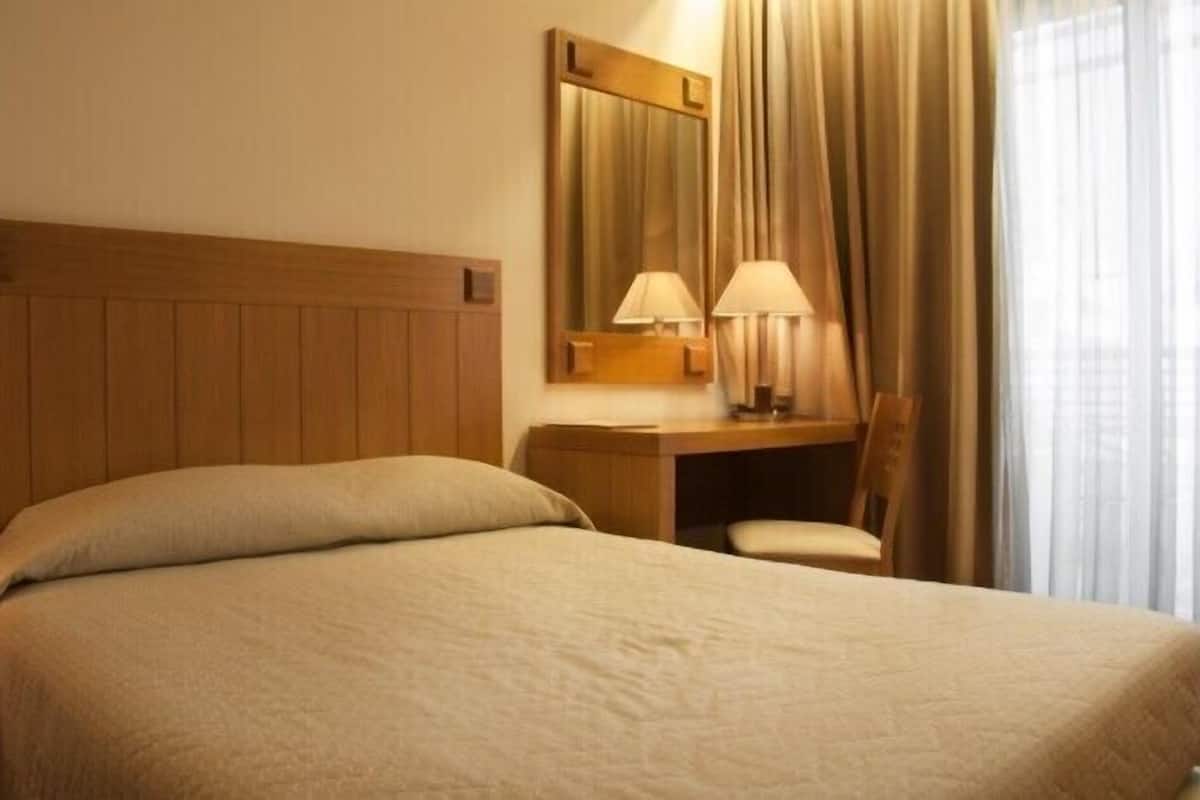 A hotel room with a bed, a desk with a chair, a lamp, and a mirror.