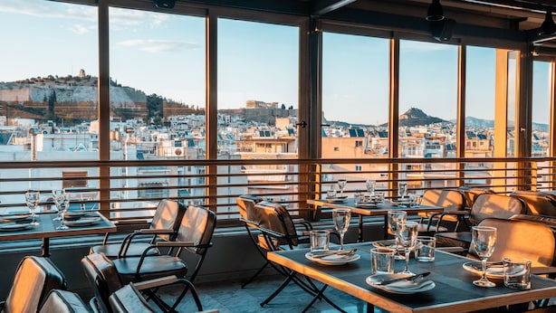 Un restaurante con amplios ventanales que ofrecen vistas de la ciudad, incluyendo una colina con ruinas. Las mesas están preparadas con platos, vasos y cubiertos.