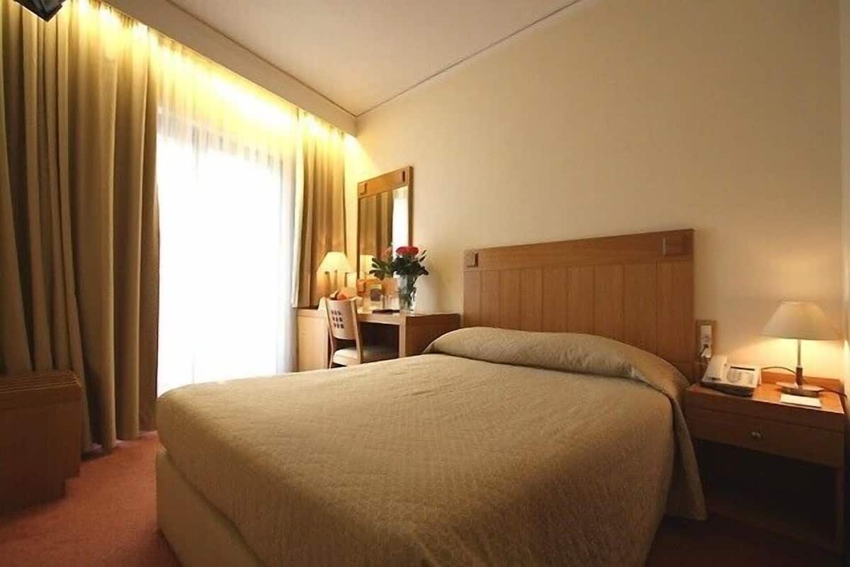 A hotel room with a large bed, a desk with a chair, a nightstand with a lamp, and a window with curtains.