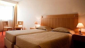 Habitación de hotel con dos camas, cabecera de madera, mesita de noche con lámpara, una silla y una pequeña mesa.