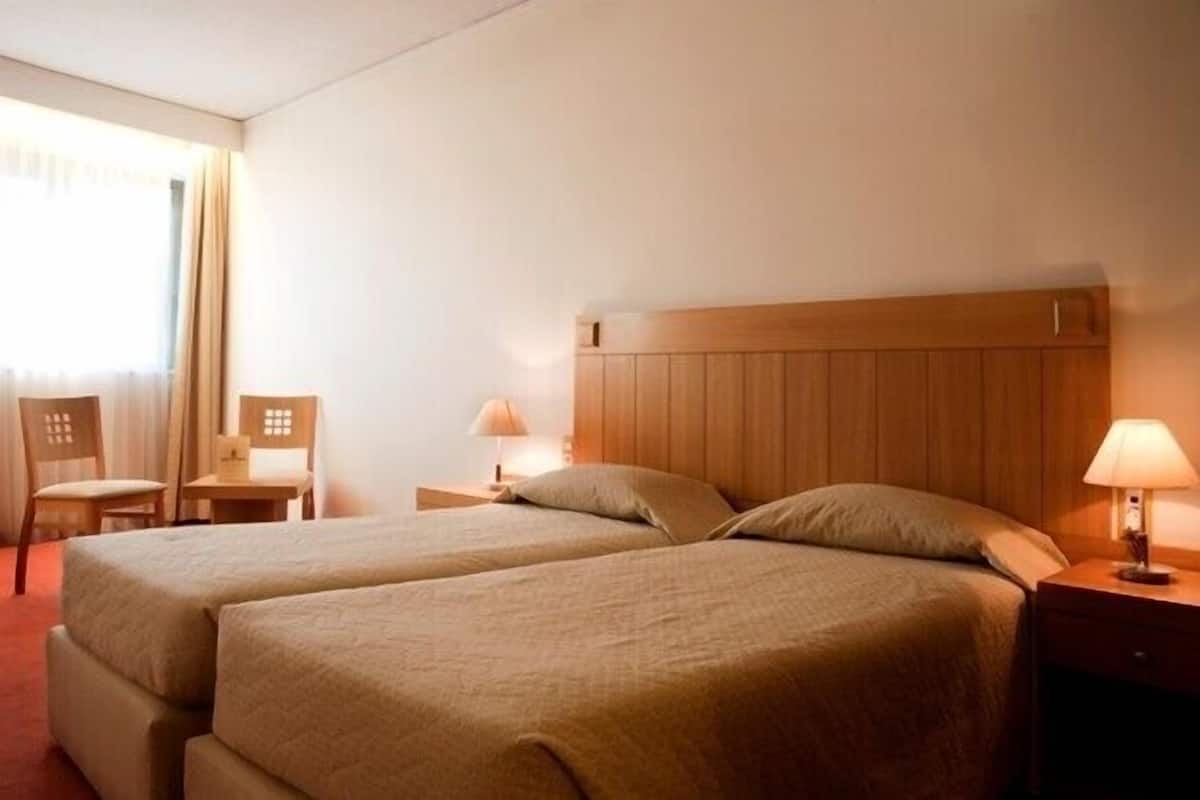 A hotel room with two beds, a wooden headboard, a nightstand with a lamp, a chair, and a small table.