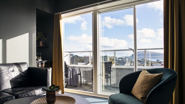 Een moderne woonkamer met een zwarte leren bank, een ronde tafel met een plant en een groene fauteuil met een kussen. Een grote schuifglazen deur leidt naar een buitenbalkon met uitzicht op gebouwen en een berg.