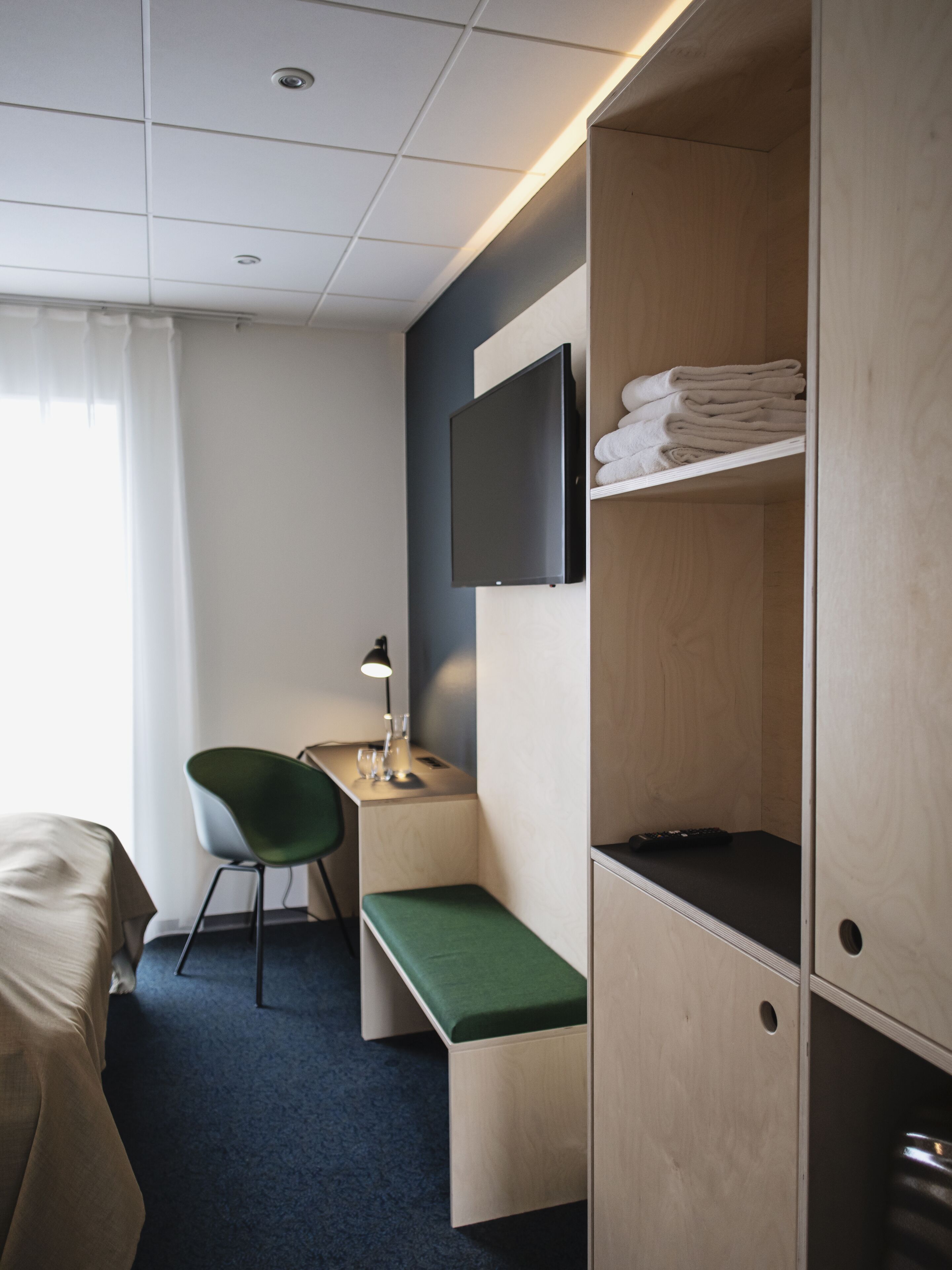 A hotel room with a bed, a desk with a chair, a TV mounted on the wall, and a wardrobe with a shelf holding folded towels.