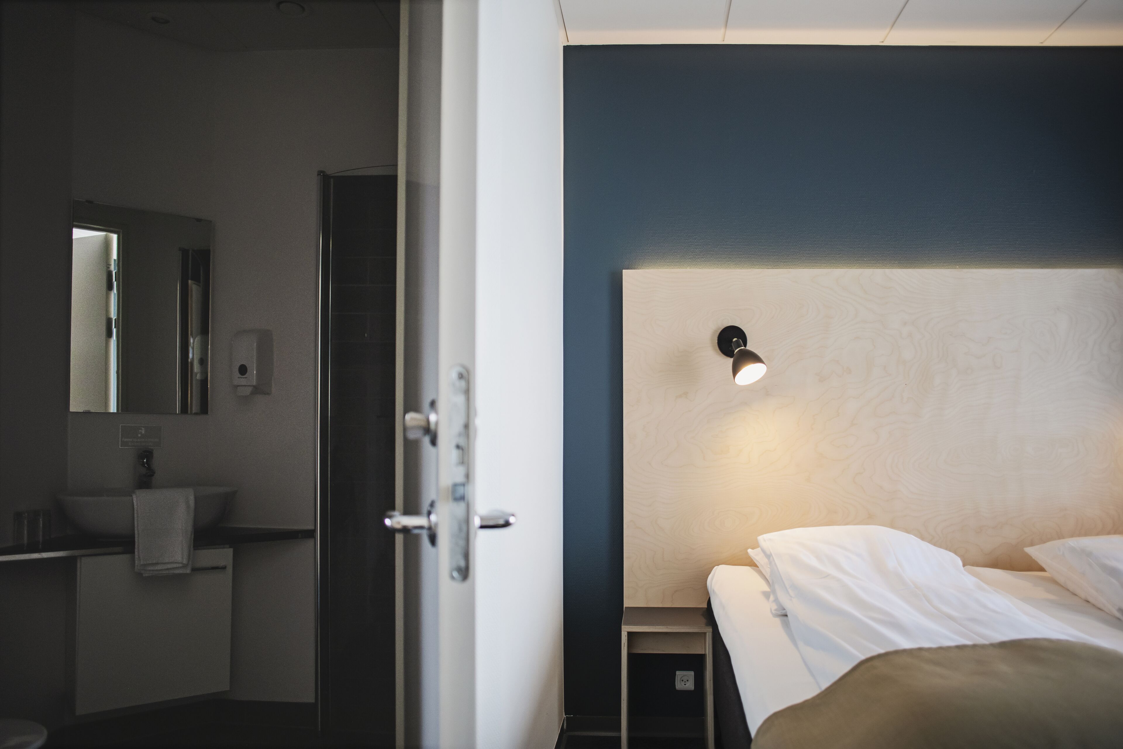 A hotel room with a bed, a bedside table, a nightstand, a mirror, a sink, and a towel.