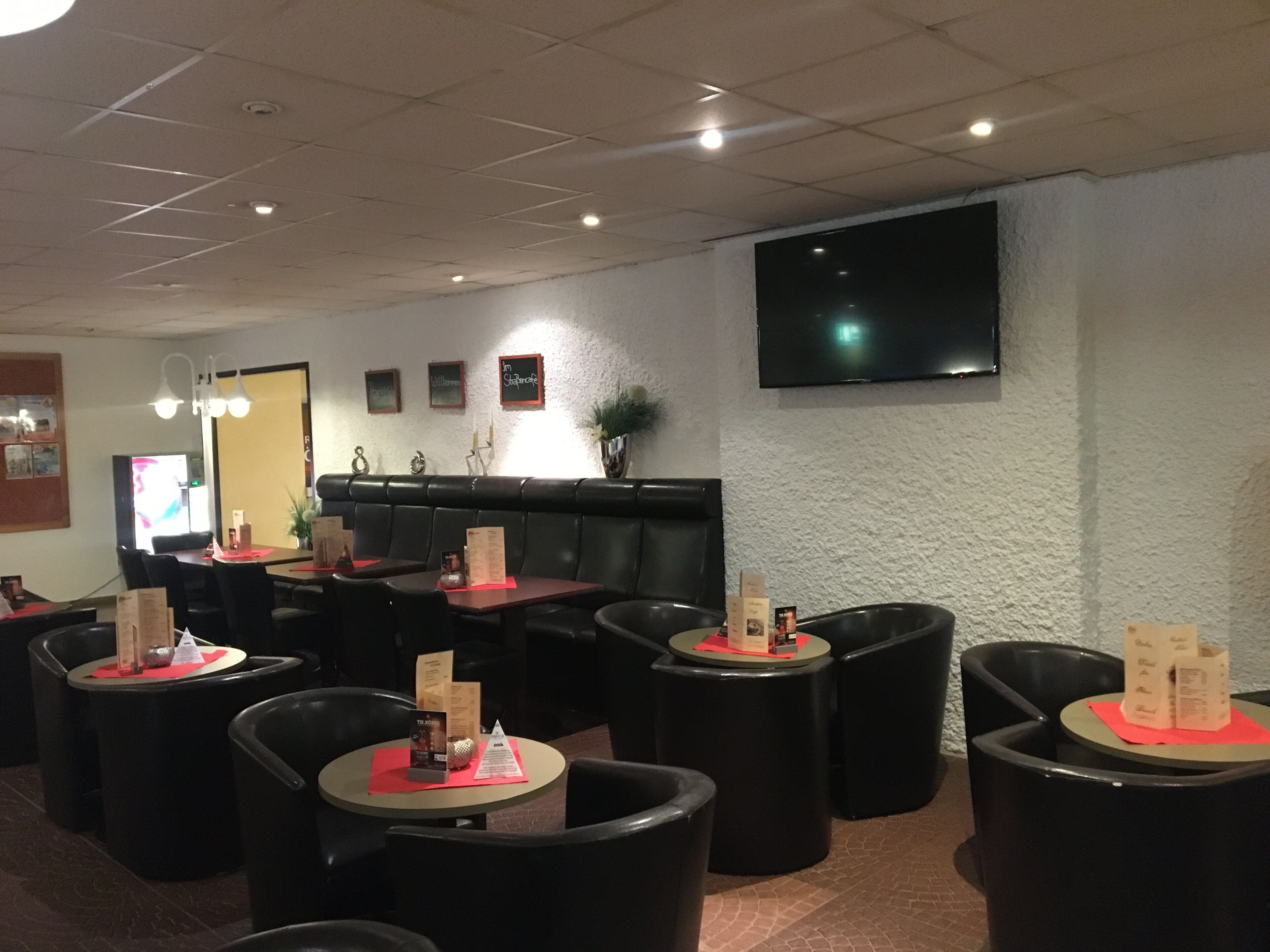 A restaurant with round tables and black chairs, a high ceiling with recessed lighting, and a flat-screen TV mounted on the wall.