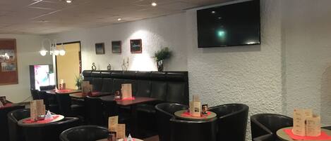 En restaurang med runda bord och svarta stolar, ett högt tak med infälld belysning och en platt-TV som är monterad på väggen.