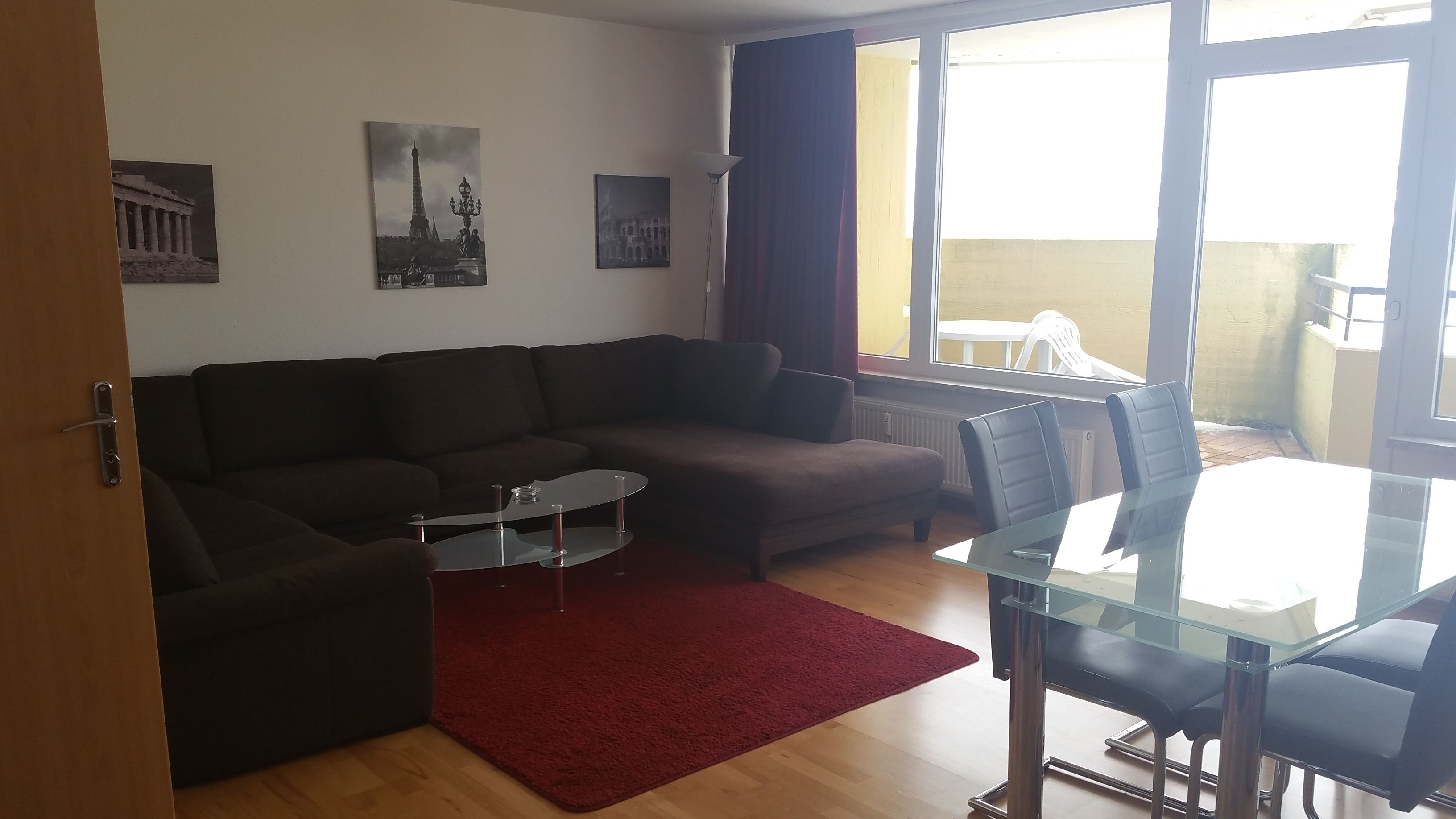 Una sala de estar con un sofá marrón, una mesa de centro de vidrio, un tapete rojo y una mesa de comedor de vidrio con sillas. Hay dos cuadros colgados en la pared, uno de la Torre Eiffel y otro de un edificio con columnas.