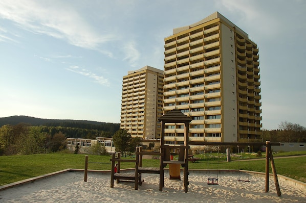 Un parque infantil con columpios y una estructura de madera, rodeado de arena, con dos edificios altos al fondo.