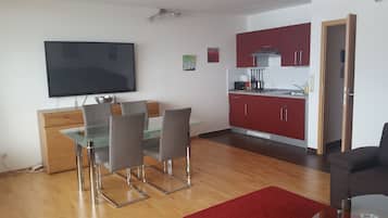 A modern living space with a dining area, a kitchen with red cabinets, a flat-screen TV, and a red rug.