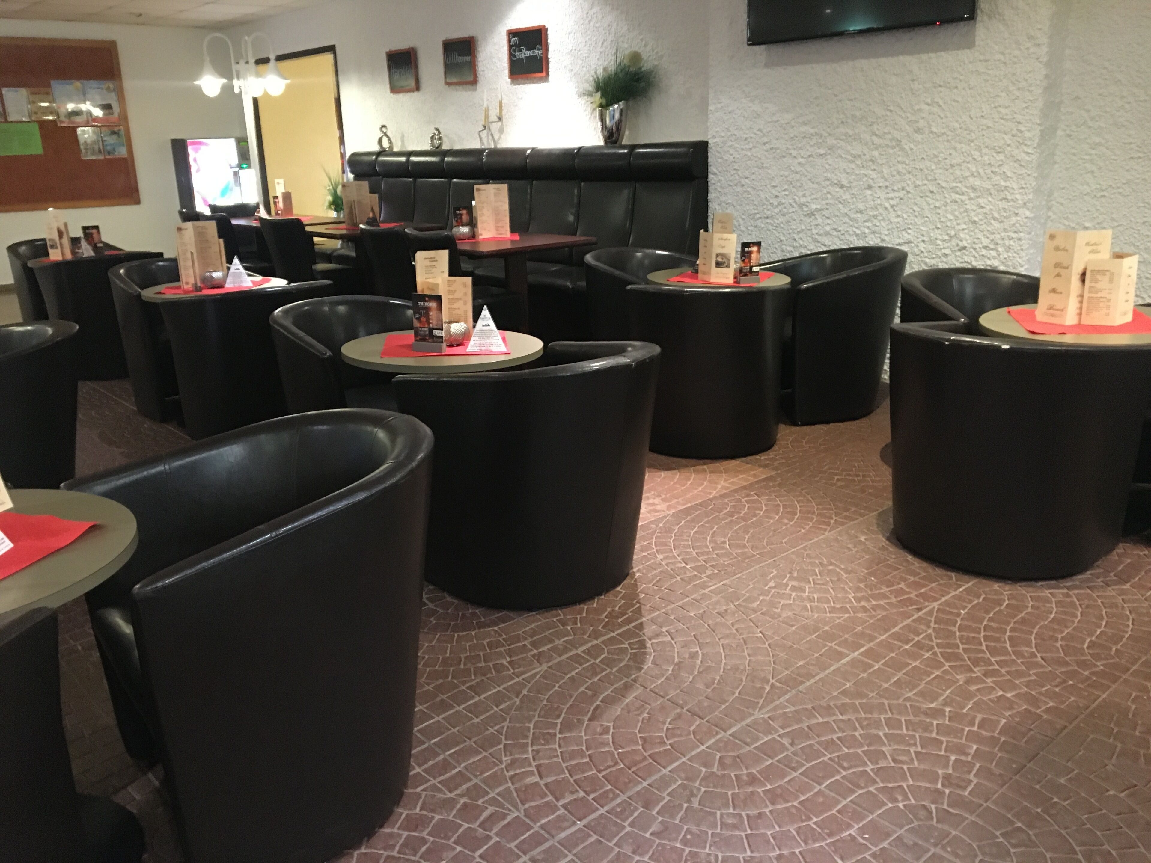 Un restaurant avec des tables rondes et des chaises noires, un sol en briques et un téléviseur à écran plat accroché au mur.
