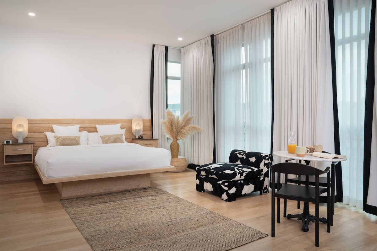 Ein modernes Hotelzimmer mit einem großen Bett, einem Essbereich mit Tisch und Stühlen sowie Blick auf die Stadt durch große Fenster.