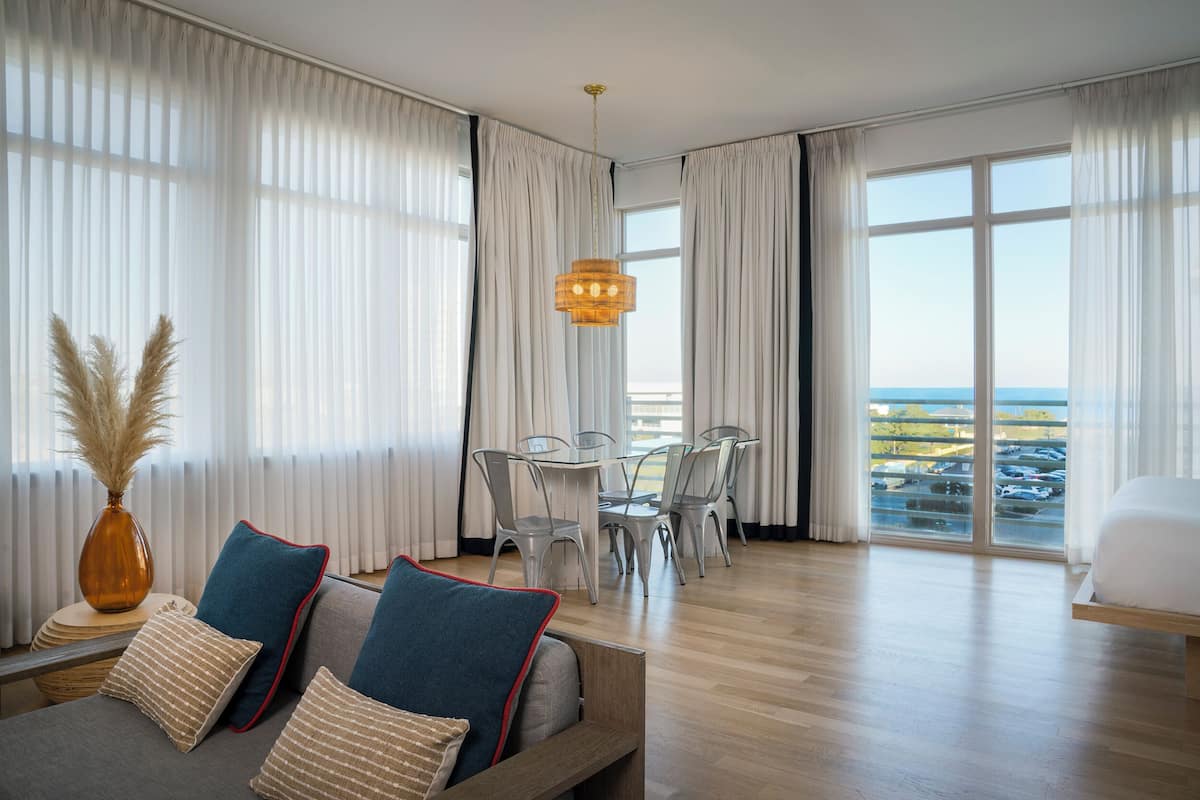 Ein modernes Hotelzimmer mit großem Fenster, einem Essbereich mit Tisch und Stühlen sowie einer Couch mit dekorativen Kissen.