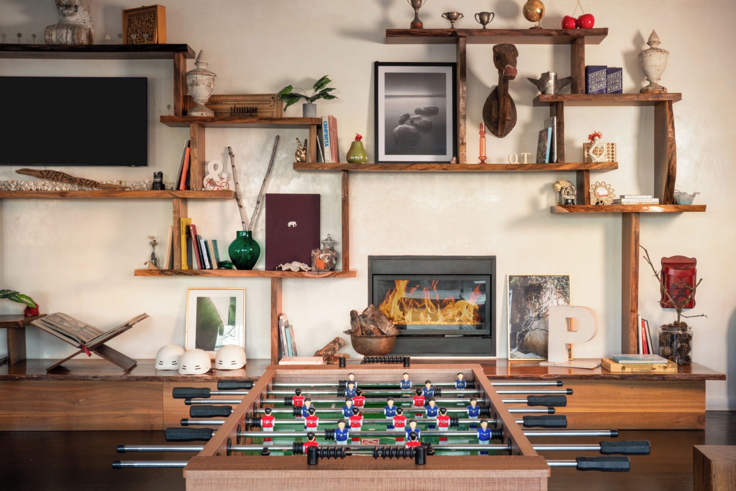 Een gezellige woonkamer met een tafelvoetbaltafel, boekenplanken vol boeken en decoratieve objecten, een open haard en een televisie.