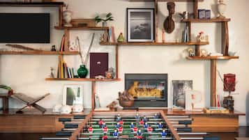 Een gezellige woonkamer met een tafelvoetbaltafel, boekenplanken vol boeken en decoratieve objecten, een open haard en een televisie.
