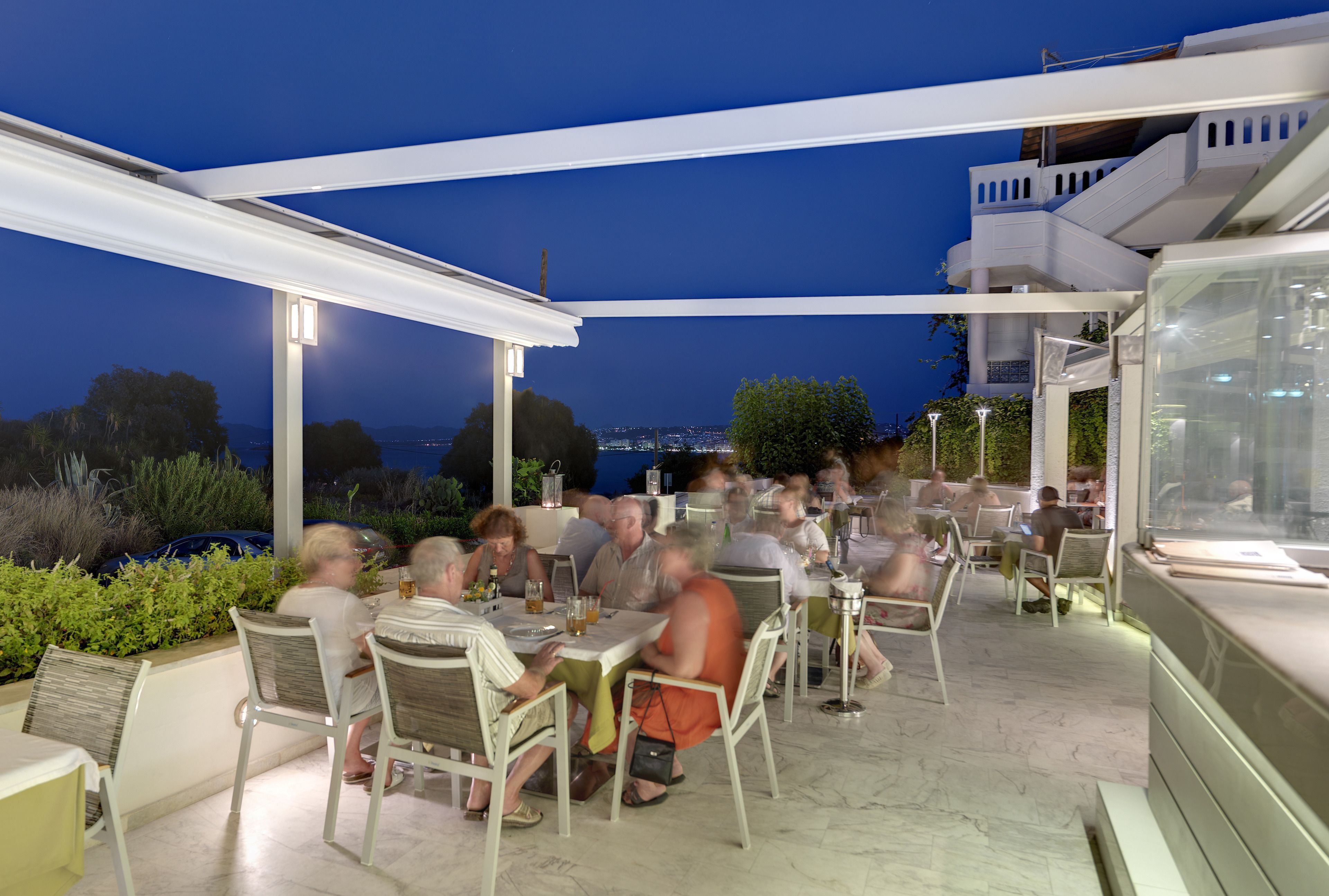 An outdoor dining area with tables and chairs, a covered section, and a view of the cityscape at dusk.