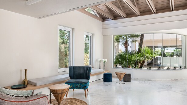 A spacious living area with a high ceiling, large windows, and a view of greenery outside.