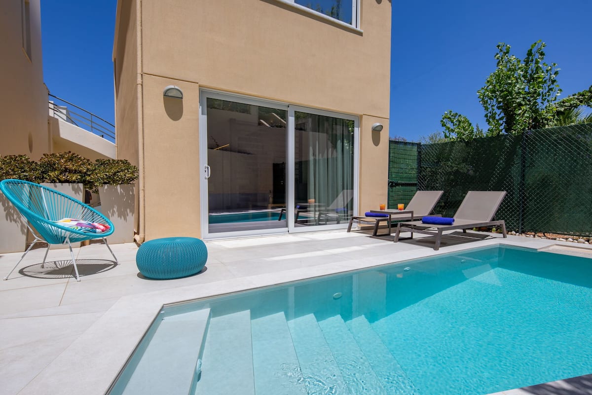 A modern outdoor pool area with a clear blue pool, two lounge chairs, a blue bean bag, and a glass door leading to a covered patio.