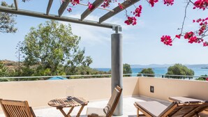 A balcony with wooden furniture, a view of the sea, and a pergola with pink flowers.